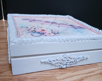 May include: White wooden box with a hinged lid. The lid features a watercolor design of pink and purple umbrellas with flowers and a pearl trim. The box has a decorative white floral design on the front.