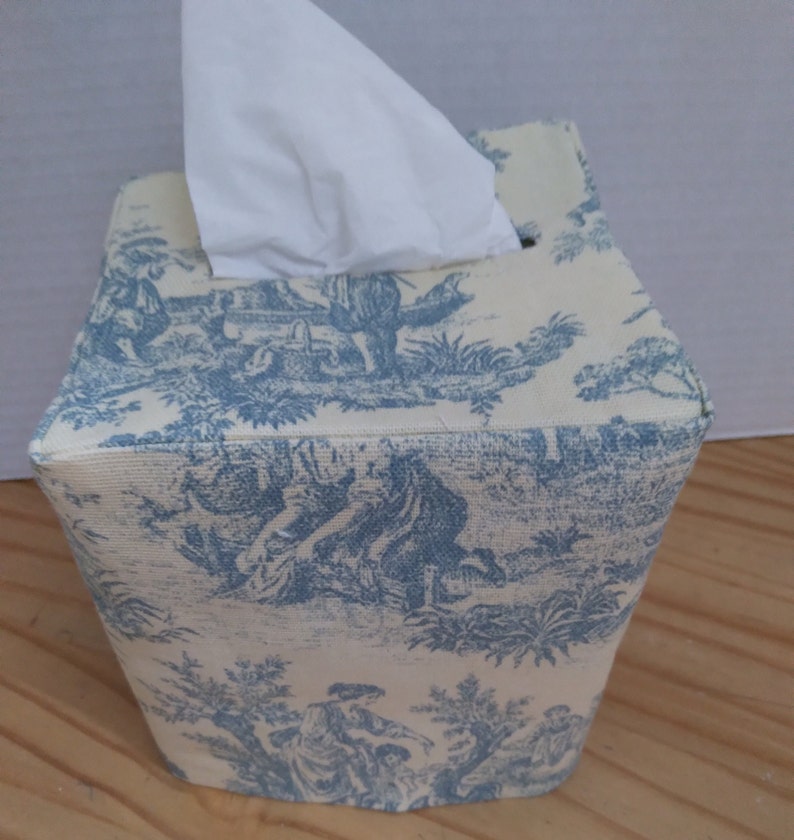 May include: A white tissue box cover with a blue and white toile pattern. The cover is square and has a small opening on the top for tissues.