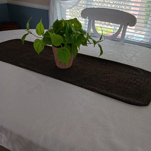 May include: A vibrant green plant in a woven basket sits on a brown leopard-print table runner. The runner is placed on a white tablecloth. A white chair and window with blinds are in the background.