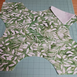May include: A green fabric with a white leaf pattern, cut into a shape for a garment. The fabric is laid out on a green cutting mat.