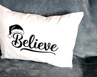 May include: White rectangular pillow with the word "Believe" in a cursive font, topped with a Santa hat graphic. The pillow is resting on a gray textured surface, creating a festive holiday theme.