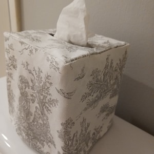 May include: A white tissue box cover with a grey toile pattern. The cover is square and has a tissue box opening on the top.