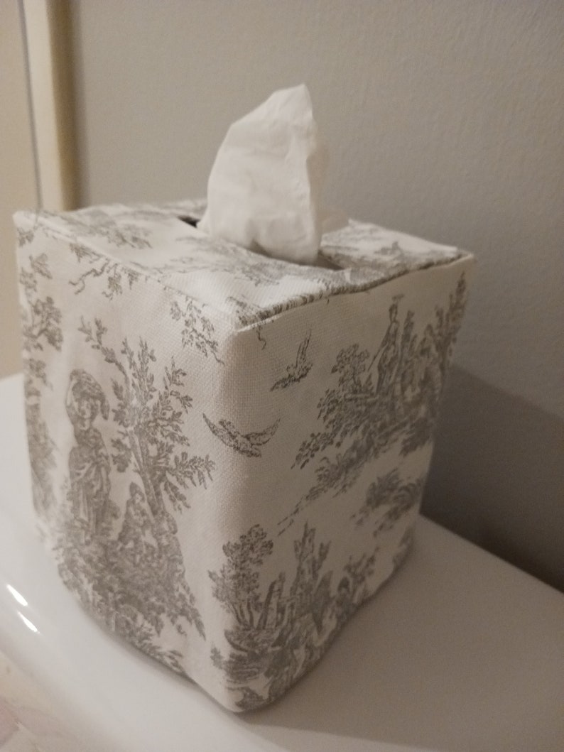 May include: A white tissue box cover with a grey toile pattern. The cover is square and has a tissue box opening on the top.