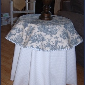 May include: A white table with a blue and white floral tablecloth. The tablecloth has a white trim around the edge. The table is in a living room setting with a lamp and a rocking chair in the background.