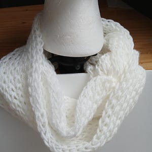May include: A white, knitted infinity scarf. The scarf is made of a soft, textured yarn and is draped around a white mannequin head. The knit pattern is open, creating a delicate and airy appearance. The scarf is a solid white color.