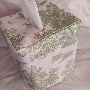 May include: A white tissue box cover with a green toile pattern. The cover is square shaped and has a tissue dispenser opening on the top.