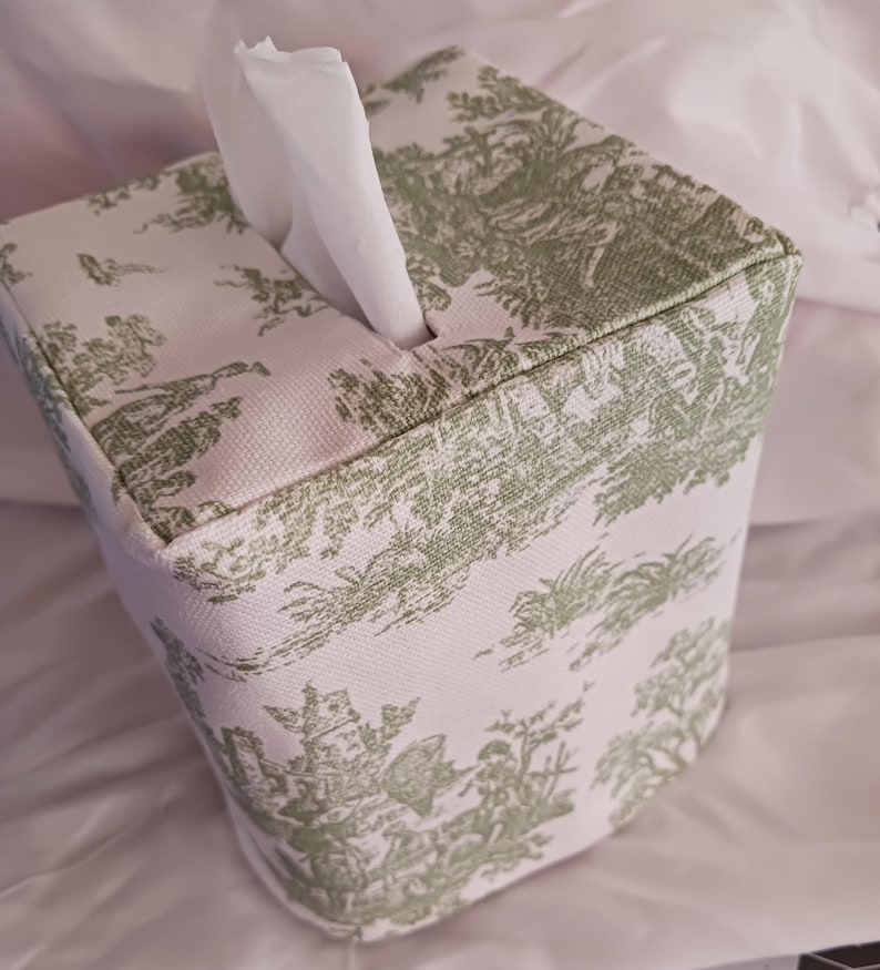 May include: A white tissue box cover with a green toile pattern. The cover is square shaped and has a tissue dispenser opening on the top.