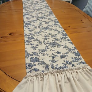 May include: A cream-colored table runner with a blue floral pattern and ruffled edges. The runner is placed on a wooden table. The floral design is a classic toile pattern, adding a touch of elegance to the table setting.