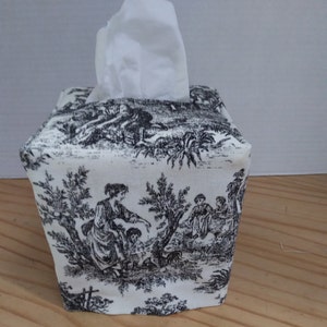 May include: A white tissue box cover with a black and white toile pattern featuring a pastoral scene with people and trees. The cover is square and has a flap on the top for pulling out tissues.