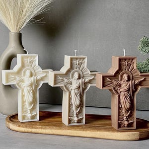 Puede incluir: Tres velas con forma de cruz en blanco, beige y marrón. Cada vela presenta un relieve de una figura con los brazos extendidos, un halo y una paloma en la parte superior. Las velas tienen una mecha blanca en el centro y una base plana.