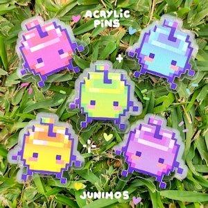 May include: Five colorful acrylic pins featuring pixelated creatures in shades of pink, blue, green, yellow, and purple. The pins have a clear border and are scattered on a bed of green grass. The text "ACRYLIC PINS" and "JUNIMOS" are visible.