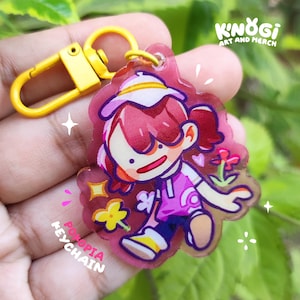 May include: A clear acrylic keychain featuring a cartoon character with pink hair and a pink outfit. The keychain has a yellow clasp and the text "POPOPIA KEYCHAIN" is visible. The brand name "KINOGI ART AND MERCH" is also visible.