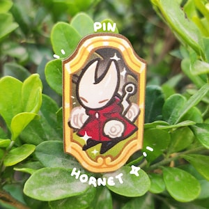 K&ouml;nnte beinhalten: Rechteckiger Ansteck-Pin mit einer Cartoon-Figur in einem roten Umhang, umrahmt von Gold. Der Pin tr&auml;gt die Worte "PIN" oben und "HORNET" unten. Der Hintergrund besteht aus gr&uuml;nen Bl&auml;ttern.