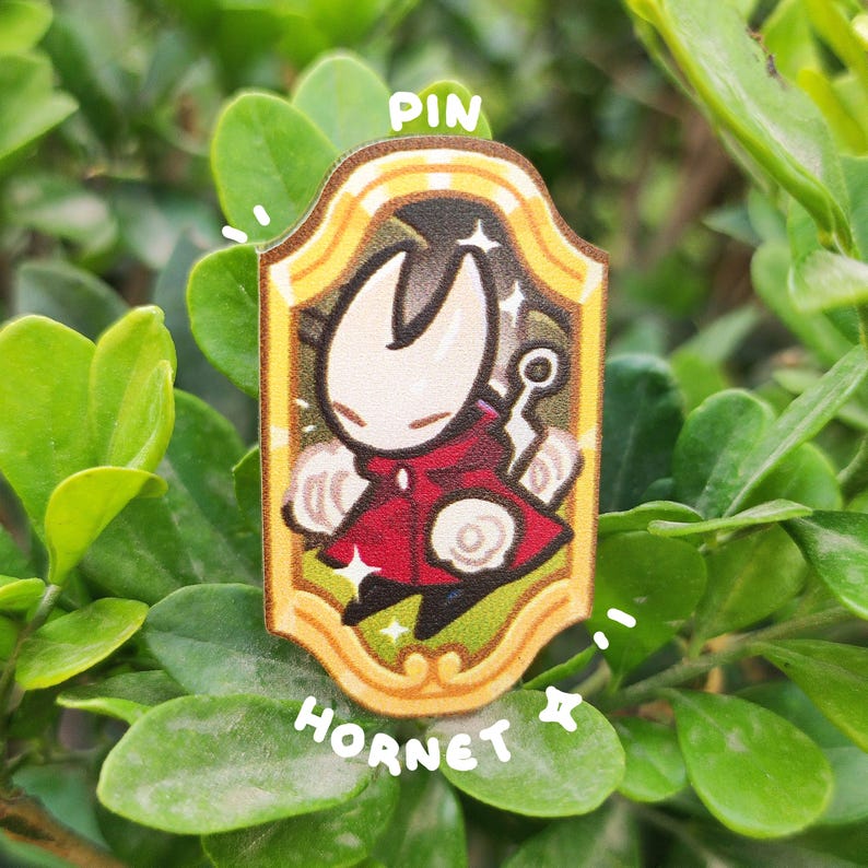 K&ouml;nnte beinhalten: Rechteckiger Ansteck-Pin mit einer Cartoon-Figur in einem roten Umhang, umrahmt von Gold. Der Pin tr&auml;gt die Worte "PIN" oben und "HORNET" unten. Der Hintergrund besteht aus gr&uuml;nen Bl&auml;ttern.
