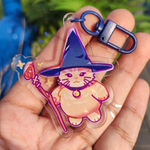 以下が含まれることがあります： 青い帽子をかぶり、杖を持った漫画の猫の魔法使いが描かれた、透明なアクリルキーホルダー。 猫は薄いオレンジ色で、ピンクのアクセントが付いています。 キーホルダーには青い留め金が付いています。