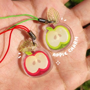 Könnte beinhalten: Zwei Apfelanhänger, einer rot und einer grün, mit einem Blatt und dem Text "APPLE CHARM". Die Anhänger sind transparent mit weißem Innenraum und roter oder grüner Umrandung. Jeder Anhänger ist an einer roten oder grünen Kordel befestigt.