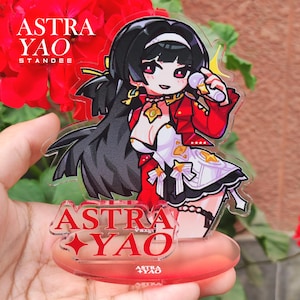 May include: A clear acrylic standee featuring a cartoon character with black hair, wearing a red jacket and white dress. The standee has the text "ASTRA YAO" in red. The character is holding a microphone. The standee is on a red base.