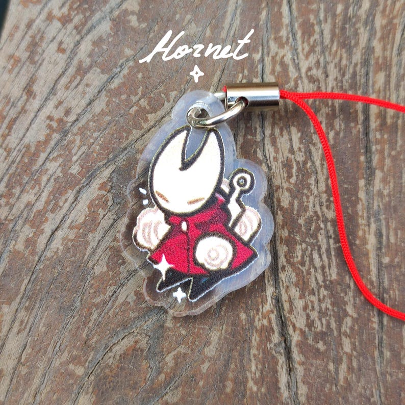 May include: A clear acrylic charm featuring a character in a red cloak with a white face and a silver clasp. The charm is attached to a red cord. The word "Hornet" is written in white script at the top.