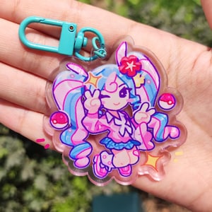 May include: A clear acrylic keyring featuring a cartoon character with blue and pink hair, wearing a pink outfit and a blue skirt. The keyring has a teal clasp and two pink and white ball-shaped charms. The character is winking and making a peace sign.