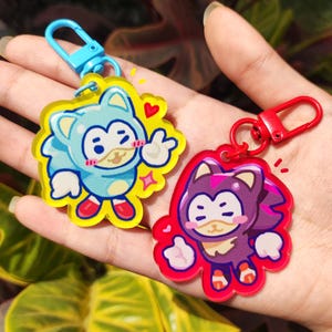 May include: Two acrylic keychains featuring cartoon characters. One is light blue with a blue clasp, the other is red with a red clasp. Both keychains have a yellow outline and a cartoon character giving a peace sign.