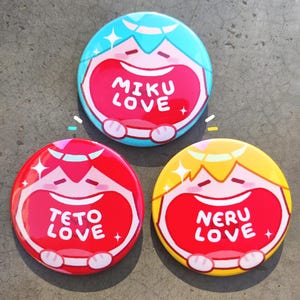 May include: Three round, colorful buttons with cartoon character designs. The buttons feature a smiling face with open mouths and the words "MIKU LOVE", "TETO LOVE", and "NERU LOVE" in white text. The buttons have blue, red, and yellow accents.