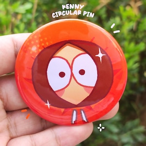 May include: A round pin with a cartoon character design, showing an orange hooded figure with a visible face. The pin has the text "KENNY CIRCULAR PIN" at the top. The pin is held in a hand against a blurred green background.