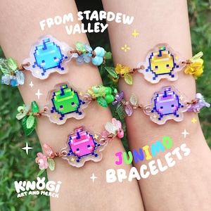 Pode incluir: Seis pulseiras Junimo com pingentes de personagens pixelados em azul, amarelo, verde, rosa e roxo. Cada pulseira apresenta um design floral com folhas e flores coloridas. O texto na imagem diz "FROM STARDEW VALLEY" e "JUNIMO BRACELETS + K'NOGI ART AND MERCH."