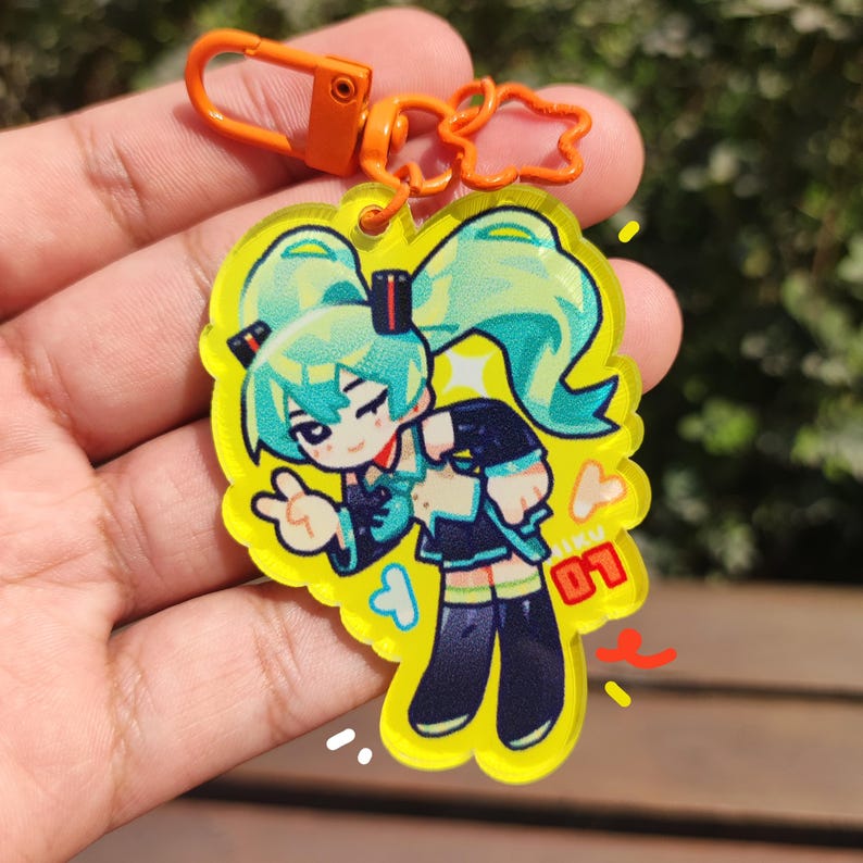May include: A yellow acrylic keychain featuring a cartoon character with teal pigtails, a black and teal outfit, and the number "01". The keychain has an orange clasp and a decorative orange loop.