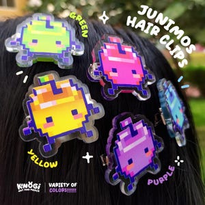 Puede incluir: Una colección de pinzas para el pelo Junimos en varios colores, incluyendo verde, rosa, amarillo, morado y azul. Cada pinza presenta un diseño pixelado y está sujeta a una pinza metálica. La imagen también incluye el texto "JUNIMOS HAIR CLIPS" y "VARIETY OF COLORS!!!!!!".