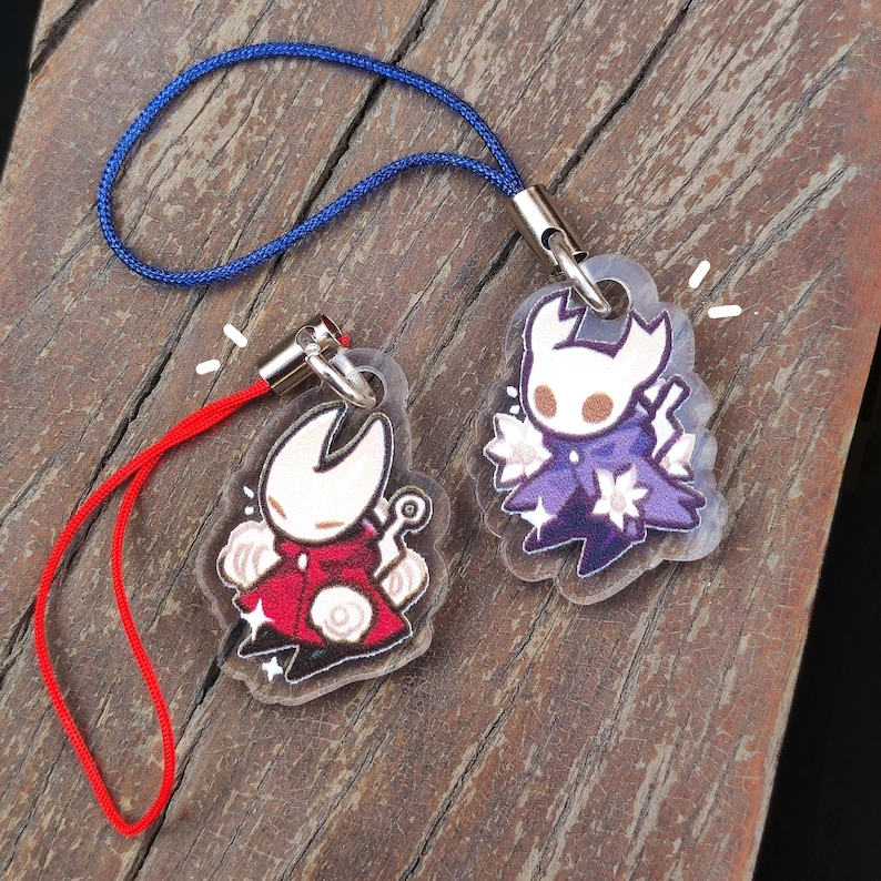 May include: Two acrylic charms featuring cartoon characters, one in red and the other in purple, each with a lanyard. The charms have a clear background and are attached to a blue and red lanyard. The charms are on a wooden surface.