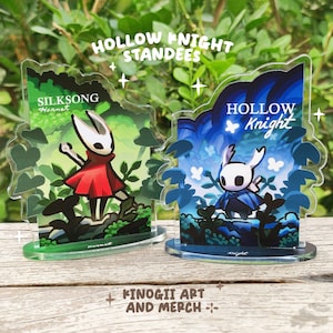 Puede incluir: Dos expositores acrílicos con personajes de Hollow Knight y Silksong. Los expositores tienen una base verde y una ilustración detallada de los personajes en un entorno forestal. El texto "HOLLOW KNIGHT STANDEES" está en la parte superior.