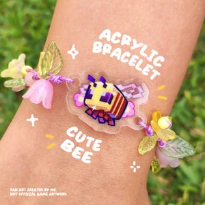 Könnte beinhalten: Ein Acrylarmband mit einem zentralen Anhänger in Form einer verpixelten gelben und braunen Biene mit violetten Augen. Das Armband ist auf eine violette Schnur gefädelt und mit durchscheinenden rosa und gelben blütenförmigen Perlen sowie grünen Blattanhängern verziert.