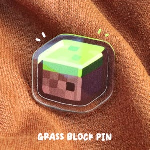 May include: A clear acrylic pin featuring a square grass block design with a bright green top and brown pixelated sides. The pin has a smooth, glossy finish and is attached to a textured orange fabric. The text GRASS BLOCK PIN is visible.