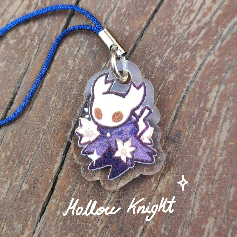 May include: A clear acrylic charm featuring a character from Hollow Knight, with a white head, purple cape, and floral accents. The charm is attached to a blue cord. The text "Hollow Knight" is written in a cursive font.