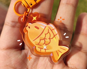 Könnte beinhalten: Ein klarer Acryl-Schlüsselanhänger mit einem cartoonartigen, fischförmigen Gebäck in Orange- und Gelbtönen. Er hat einen goldfarbenen Verschluss und Ring. Das Design umfasst Stern- und Kreuzakzente.