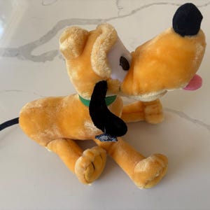 May include: A plush toy of a yellow dog with long black ears, a black nose, and a green collar. The character has a white muzzle, a small pink tongue sticking out, and a thin black tail. A small blue tag is attached to one of the black ears.