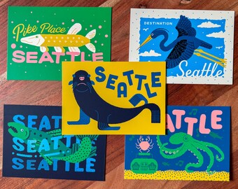 Puede incluir: Cinco postales coloridas con temática de Seattle. Los diseños incluyen un pez, una garza, un león marino, un pez y un pulpo. Cada tarjeta presenta la palabra "Seattle" en una fuente y esquema de color únicos.