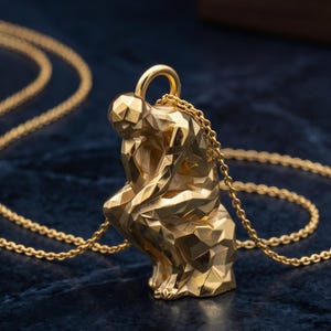 May include: A gold-toned necklace featuring a pendant shaped like a seated figure in a faceted, geometric style. The pendant is suspended from a delicate gold-toned chain, showcasing a polished metallic finish with sharp, angular surfaces.