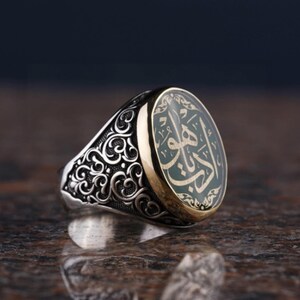 May include: A silver ring with an oval-shaped green stone set in a gold bezel. The ring features intricate floral designs around the stone.