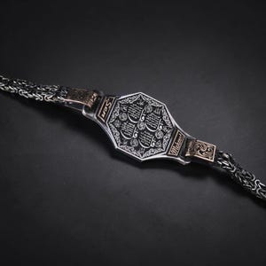 Op de afbeelding: Een zilveren armband met een achthoekig middelpunt met ingewikkelde gegraveerde patronen en Arabisch schrift. De armband heeft een kettingachtig ontwerp met goudkleurige accenten en de namen "Son" en "Mehmet" op de zijkanten. De achtergrond is donker.