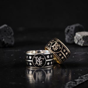 May include: Two rings with a black band and silver or gold accents. Each ring features a central emblem and maple leaf designs. The rings are displayed on a dark surface, showcasing their intricate details.