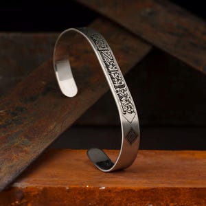 Op de afbeelding: Een zilveren manchetarmband met ingewikkelde zwart geëtste ontwerpen. De open armband heeft een geometrisch patroon en script langs de bovenkant. De sieraden worden gepresenteerd op een bruin oppervlak met een donkere achtergrond.