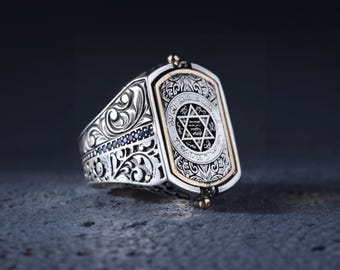 May include: A silver ring with a gold-toned rectangular center featuring a Star of David with Hebrew text. The ring is decorated with intricate floral designs and blue gemstones.
