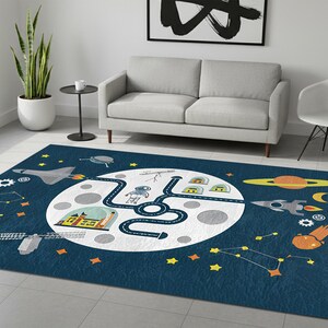 以下が含まれることがあります： 宇宙をテーマにした長方形の濃い紺色のラグ。デザインは、道路、建物、宇宙飛行士がいる大きな白い月が特徴です。ロケット、惑星、星、星座が散らばっています。