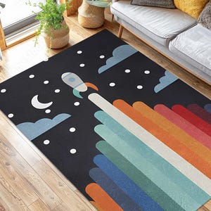 以下が含まれることがあります： 宇宙をテーマにしたデザインの長方形の黒いラグ。ロケットが三日月、雲、星のある夜空に打ち上げられます。ロケットから虹色の軌跡が伸び、床材に鮮やかなタッチを加えています。