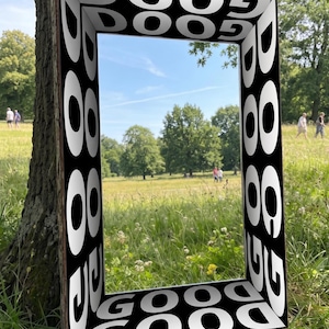 以下が含まれることがあります： 縁に「GOOD」の文字が繰り返された、長方形の白黒フレーム。 フレームは木の幹に配置され、開口部からは草、木、人々を含む公園の風景が見えます。
