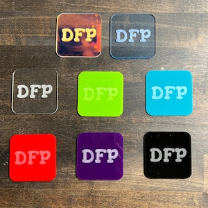 May include: A collection of square acrylic coasters in various colors, each featuring the white letters "DFP". Colors include clear, red, purple, black, blue, green, and a multi-colored iridescent option. The coasters are displayed on a dark wood surface.