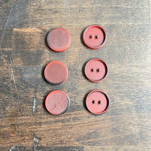 May include: Six red and black round buttons with a textured surface. The buttons have two small holes in the center.