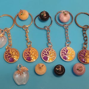 Op de afbeelding: Vijf sleutelhangers met een levensboomontwerp. Elke sleutelhanger heeft een hanger van een steen in een andere kleur: geel, zwart, roze, wit en een heldere steen. Het levensboomontwerp is paars en geel.