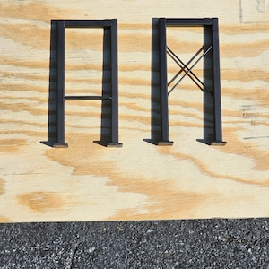 May include: Two black metal table legs, one with a simple design and the other with an X-shaped brace. The legs are on a wooden surface.